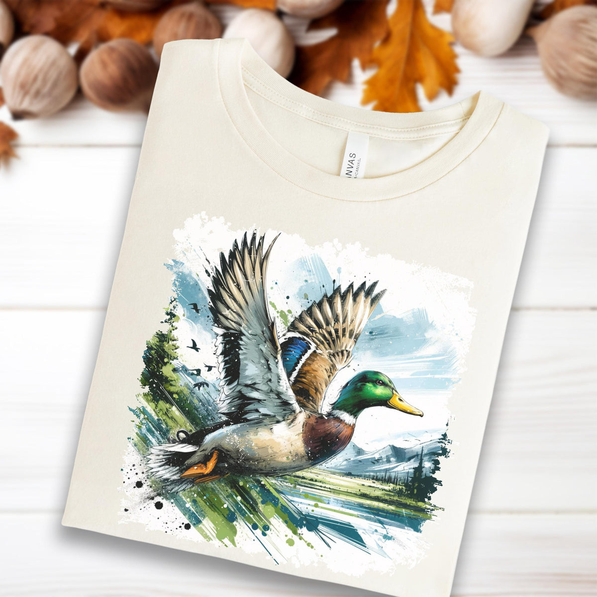May include: Cream-colored t-shirt featuring a watercolor illustration of a flying mallard duck. The duck has green, brown, and blue plumage, with wings spread wide. The background includes a landscape with trees, mountains, and a blue sky.