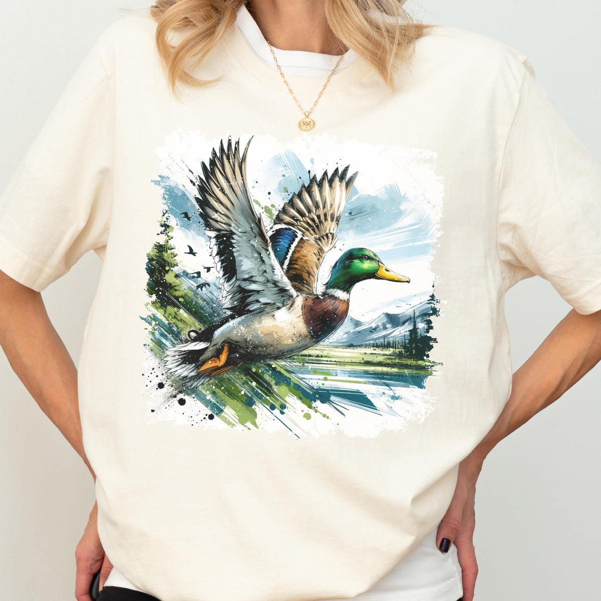 May include: Cream-colored t-shirt featuring a watercolor illustration of a flying mallard duck. The duck has green, brown, and blue feathers, with a yellow beak. The background includes a landscape with trees, mountains, and a blue sky.