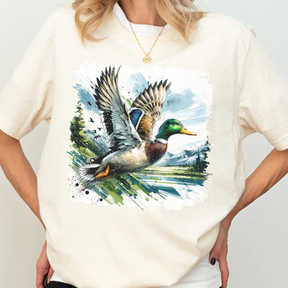 May include: Cream-colored t-shirt featuring a watercolor illustration of a flying mallard duck. The duck has green, brown, and blue feathers, with a yellow beak. The background includes a landscape with trees, mountains, and a blue sky.