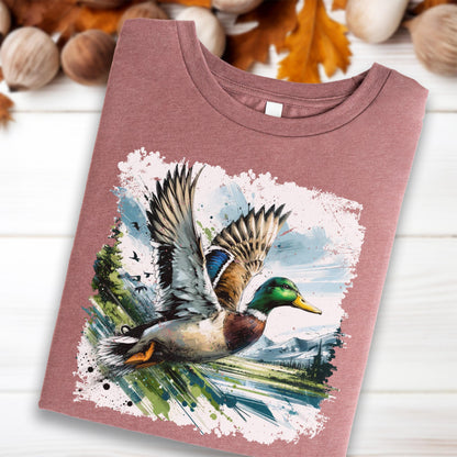 May include: A mauve-colored t-shirt featuring a watercolor painting of a mallard duck in flight. The duck has green, brown, blue, and white feathers. The background includes trees, mountains, and a blue sky.