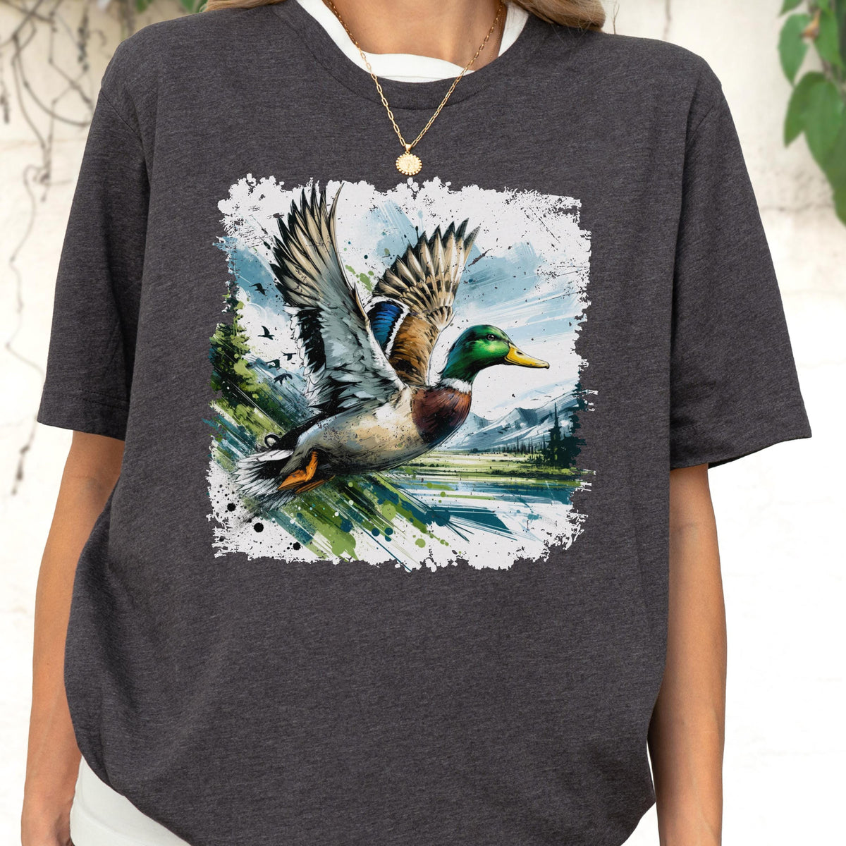 May include: Dark gray t-shirt featuring a watercolor-style image of a mallard duck in flight. The duck has green, blue, brown, and white feathers, with a yellow beak. The background includes a lake, trees, and mountains.