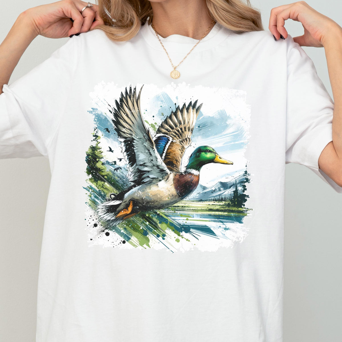May include: White t-shirt featuring a watercolor illustration of a mallard duck in flight. The duck has green, brown, and blue plumage, with a yellow beak. The background includes a landscape with trees, water, and mountains.