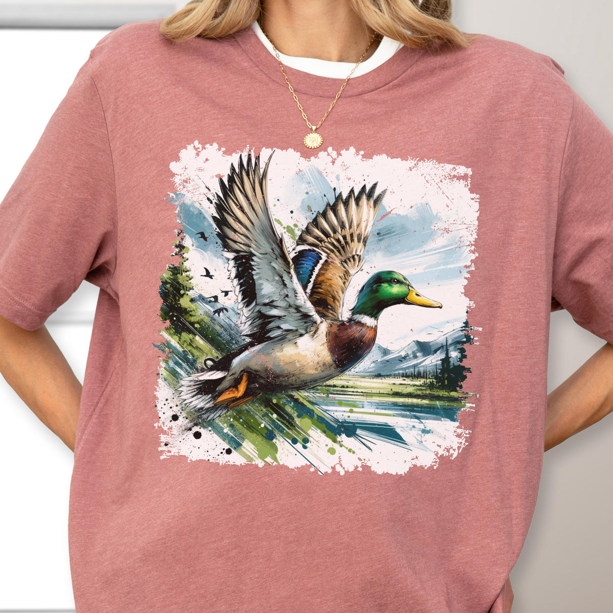 May include: A dusty rose t-shirt featuring a vibrant graphic of a mallard duck in flight. The duck has detailed feathers in shades of brown, blue, and green, set against a backdrop of a landscape with trees and mountains.