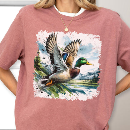 May include: A dusty rose t-shirt featuring a vibrant graphic of a mallard duck in flight. The duck has detailed feathers in shades of brown, blue, and green, set against a backdrop of a landscape with trees and mountains.
