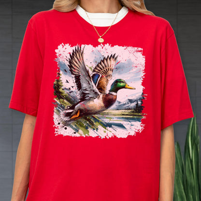 May include: Red t-shirt featuring a watercolor painting of a flying mallard duck with outstretched wings. The duck has green, brown, and blue feathers, a yellow beak, and is set against a landscape with mountains and water.