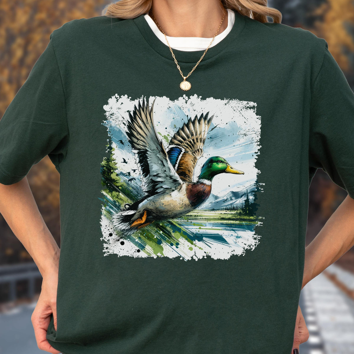 May include: Dark green t-shirt featuring a watercolor-style graphic of a mallard duck in flight. The duck has green, brown, blue, and white feathers, with a yellow beak. The background includes a landscape with trees and mountains.