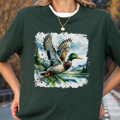 May include: Dark green t-shirt featuring a watercolor-style graphic of a mallard duck in flight. The duck has green, brown, blue, and white feathers, with a yellow beak. The background includes a landscape with trees and mountains.