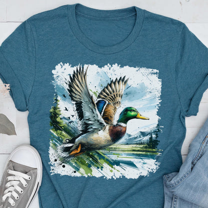 May include: Teal-colored t-shirt featuring a detailed illustration of a mallard duck in flight. The duck has green, brown, and blue plumage, with wings spread wide. The background includes a landscape with mountains and a lake.
