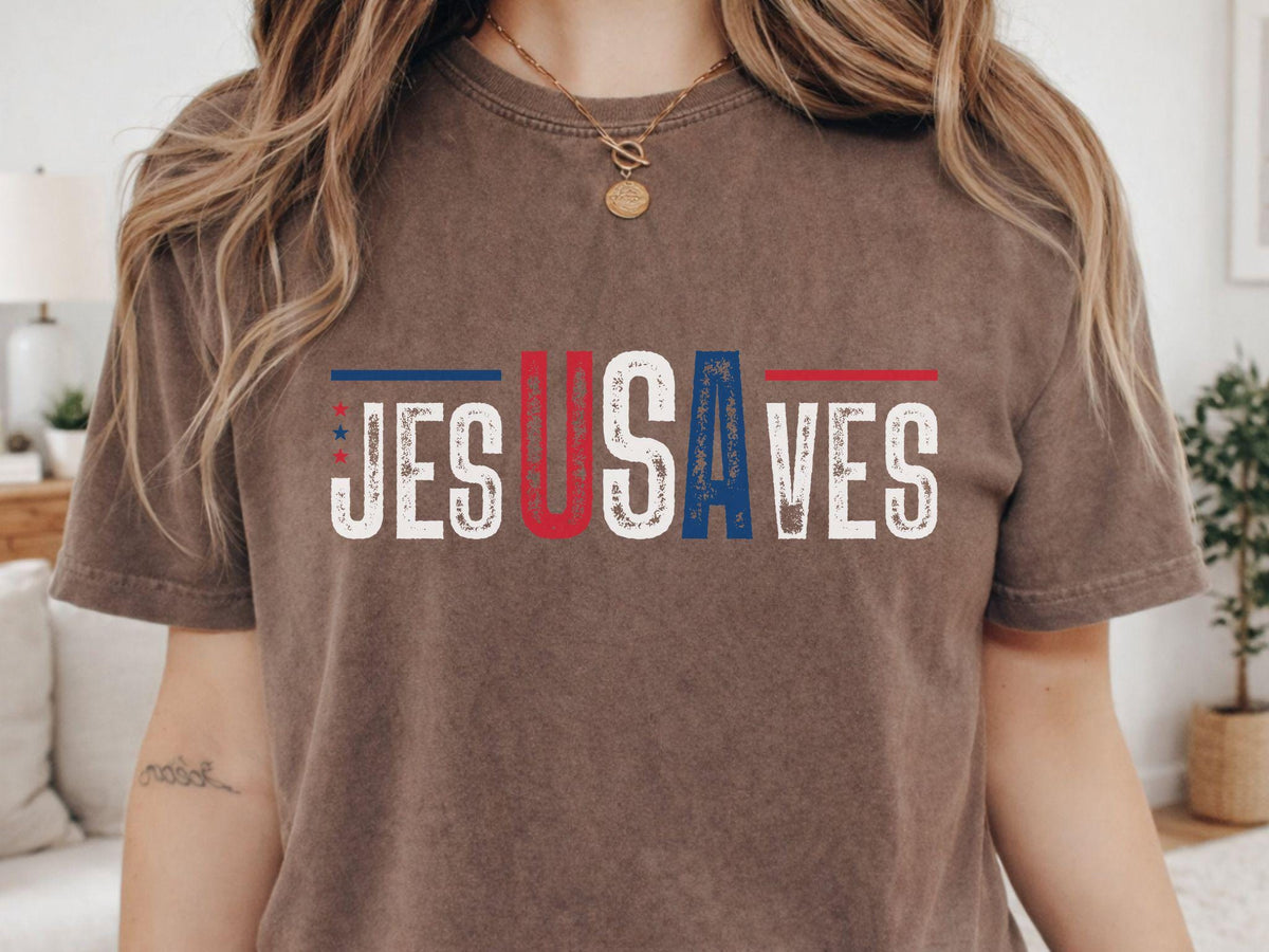 May include: A brown t-shirt with the text "JESUS SAVES" in a distressed, patriotic font. The letters are in red, white, and blue, with stars and stripes accents. The shirt has short sleeves and a crew neck.