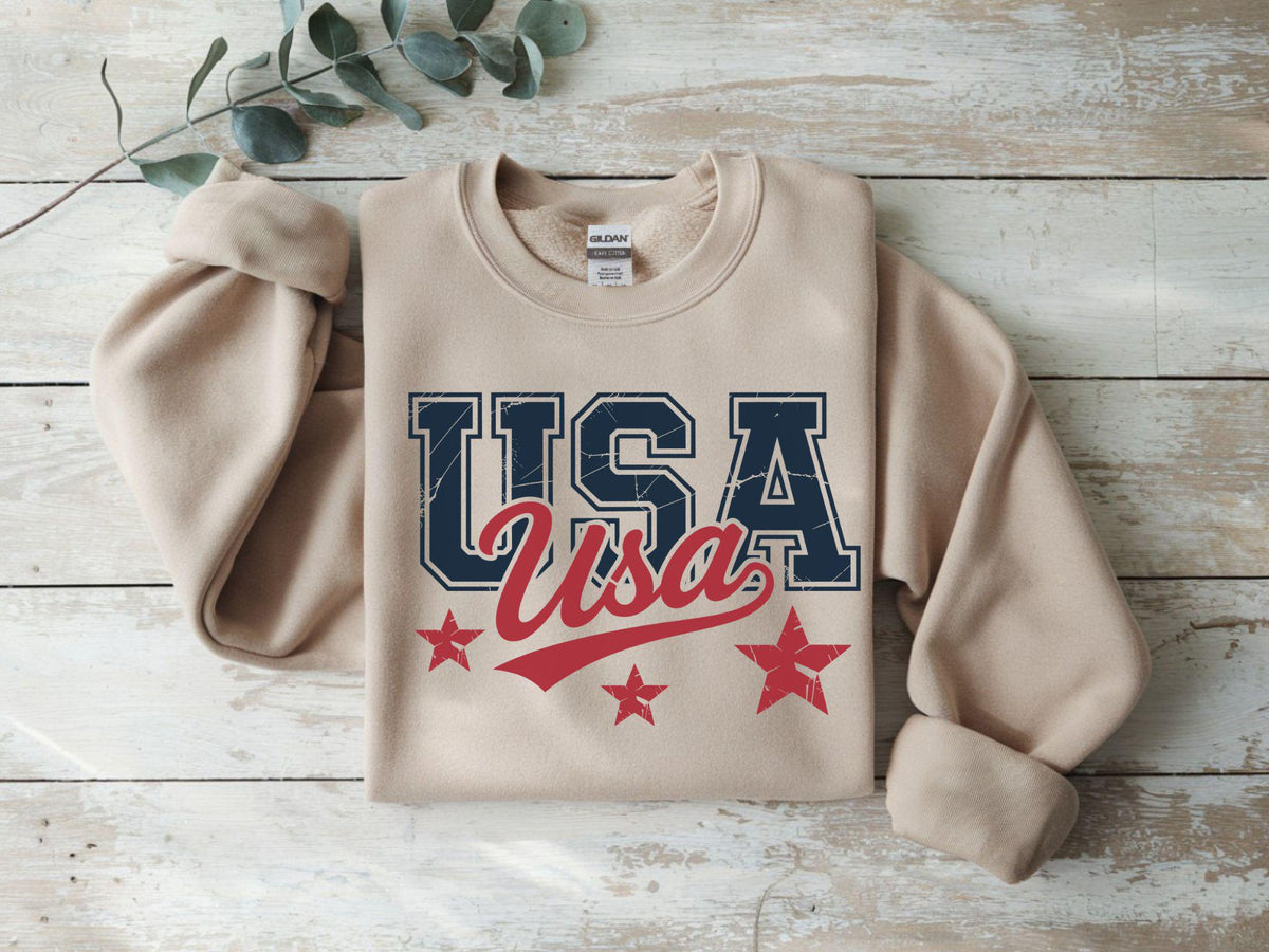 May include: A beige sweatshirt with the word "USA" in navy blue and red lettering, along with three red stars. The sweatshirt is laid flat on a light wood surface.