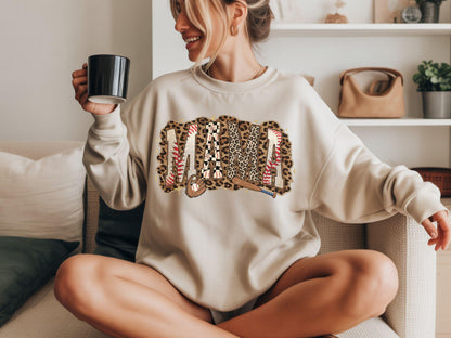 May include: Beige sweatshirt with the word "MAMA" in a baseball-themed design. The letters are decorated with leopard print, baseball stitching, and a baseball bat. The sweatshirt is a neutral color.