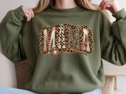 May include: Olive green sweatshirt with a baseball-themed "MAMA" graphic. The graphic features the word "MAMA" in a mix of leopard print, baseball stitching, and checkered patterns, with baseball and bat illustrations.