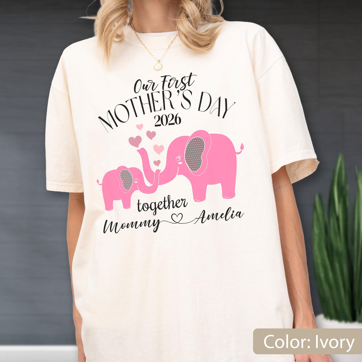 May include: Ivory-colored t-shirt with the text "Our First Mother's Day 2026" and a pink elephant design. The design includes two elephants, hearts, and the words "together Mommy Amelia".