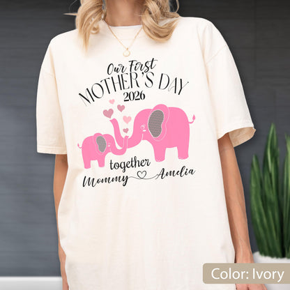 May include: Ivory-colored t-shirt with the text "Our First Mother's Day 2026" and a pink elephant design. The design includes two elephants, hearts, and the words "together Mommy Amelia".