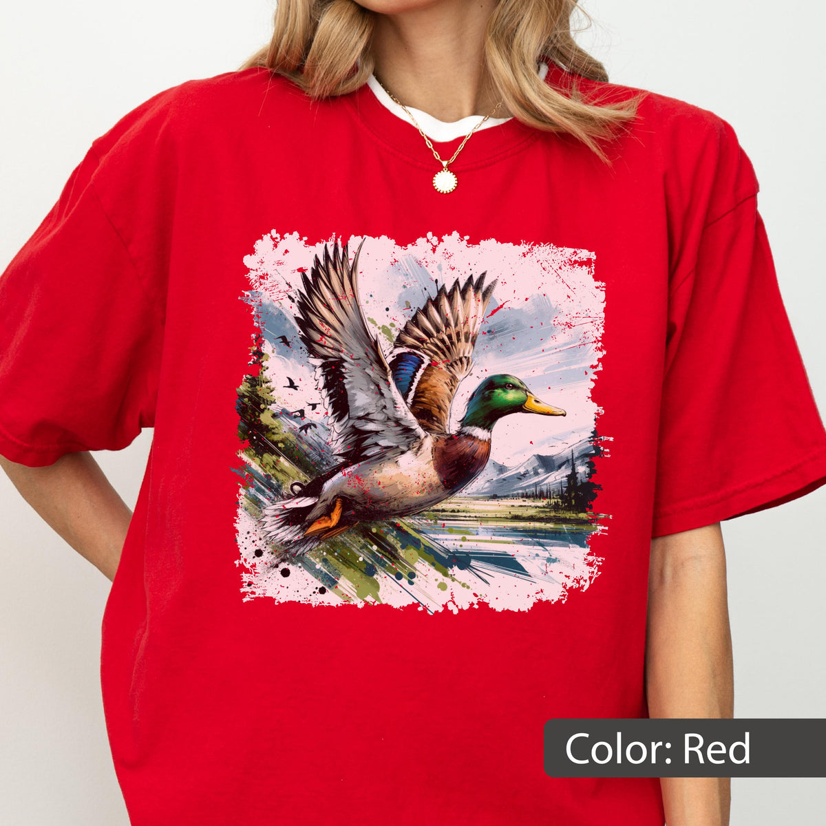 May include: Red t-shirt featuring a watercolor-style graphic of a mallard duck in flight. The duck has brown, blue, and green feathers, with a yellow beak. The background includes a landscape with trees, water, and mountains. The text "Color: Red" is at the bottom.