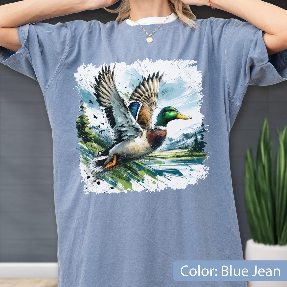 May include: Blue jean colored t-shirt featuring a watercolor painting of a mallard duck in flight. The duck has green, brown, and blue feathers. The background includes a lake, mountains, and trees. The text "Color: Blue Jean" is at the bottom.