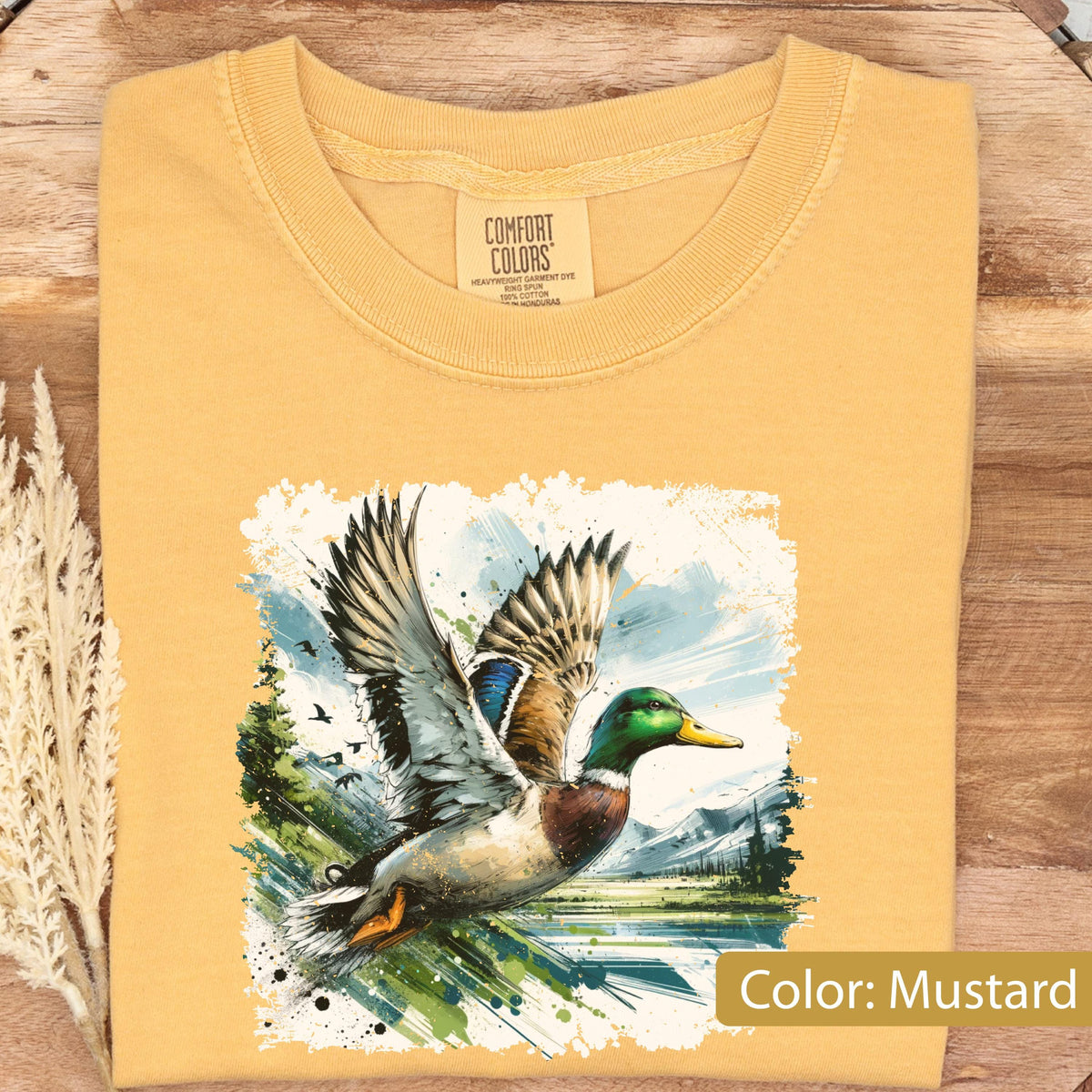 May include: Mustard-colored t-shirt featuring a detailed illustration of a duck in flight over a lake and mountains. The duck has green, blue, and brown feathers. The shirt has a crew neck and a tag that says "Comfort Colors".