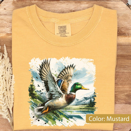 May include: Mustard-colored t-shirt featuring a detailed illustration of a duck in flight over a lake and mountains. The duck has green, blue, and brown feathers. The shirt has a crew neck and a tag that says "Comfort Colors".