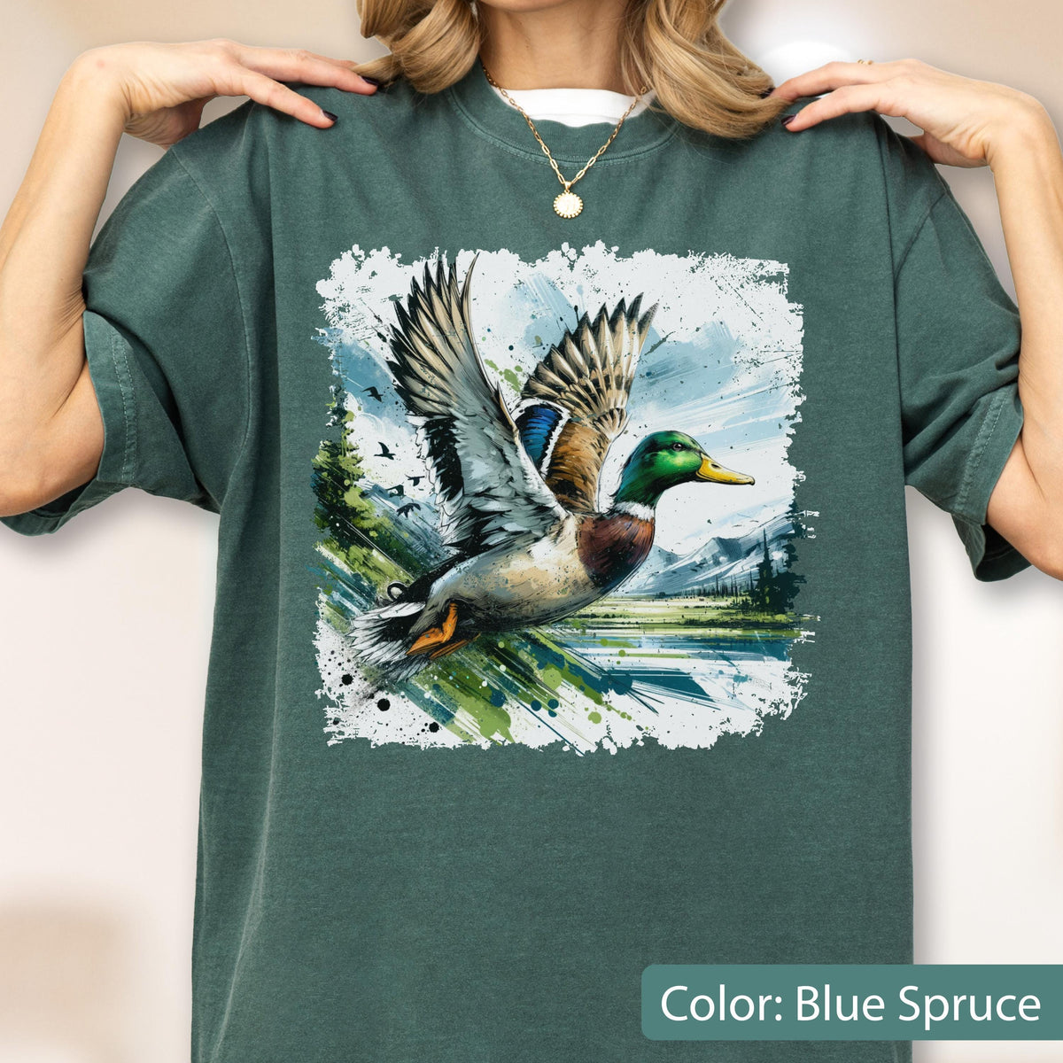 May include: Blue Spruce-colored t-shirt featuring a watercolor graphic of a mallard duck in flight. The duck has detailed feathers and is set against a landscape with trees, water, and mountains. The text "Color: Blue Spruce" is at the bottom.