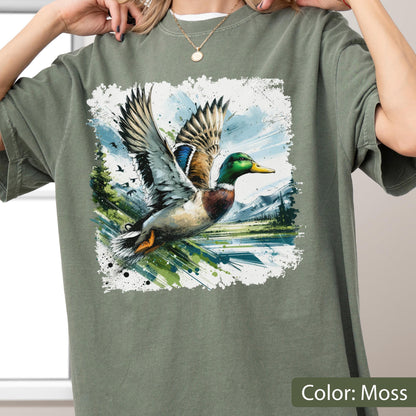 May include: Moss-colored t-shirt featuring a watercolor illustration of a mallard duck in flight. The duck has green, brown, blue, and white feathers, with a yellow beak. The background shows a lake, trees, and mountains.