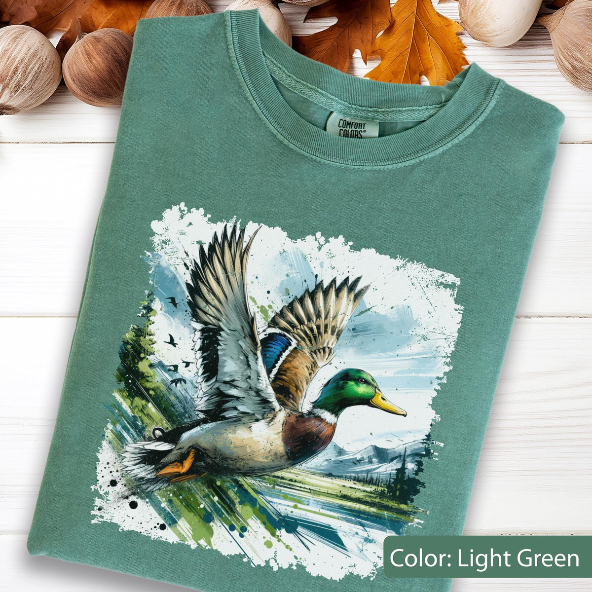 May include: Light green t-shirt featuring a watercolor-style graphic of a mallard duck in flight. The duck has detailed feathers and is set against a backdrop of a landscape with trees and mountains. The text "Color: Light Green" is at the bottom.
