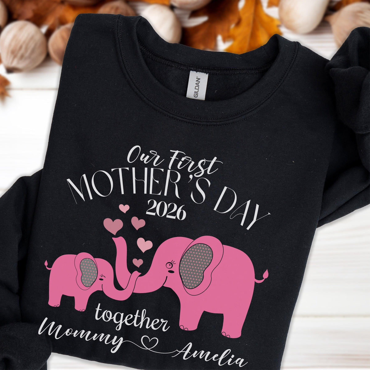 May include: Black sweatshirt with the text "Our First Mother's Day 2026" and the names "Mommy" and "Amelia". Two pink elephants are featured, with their trunks forming hearts. A heartwarming design.