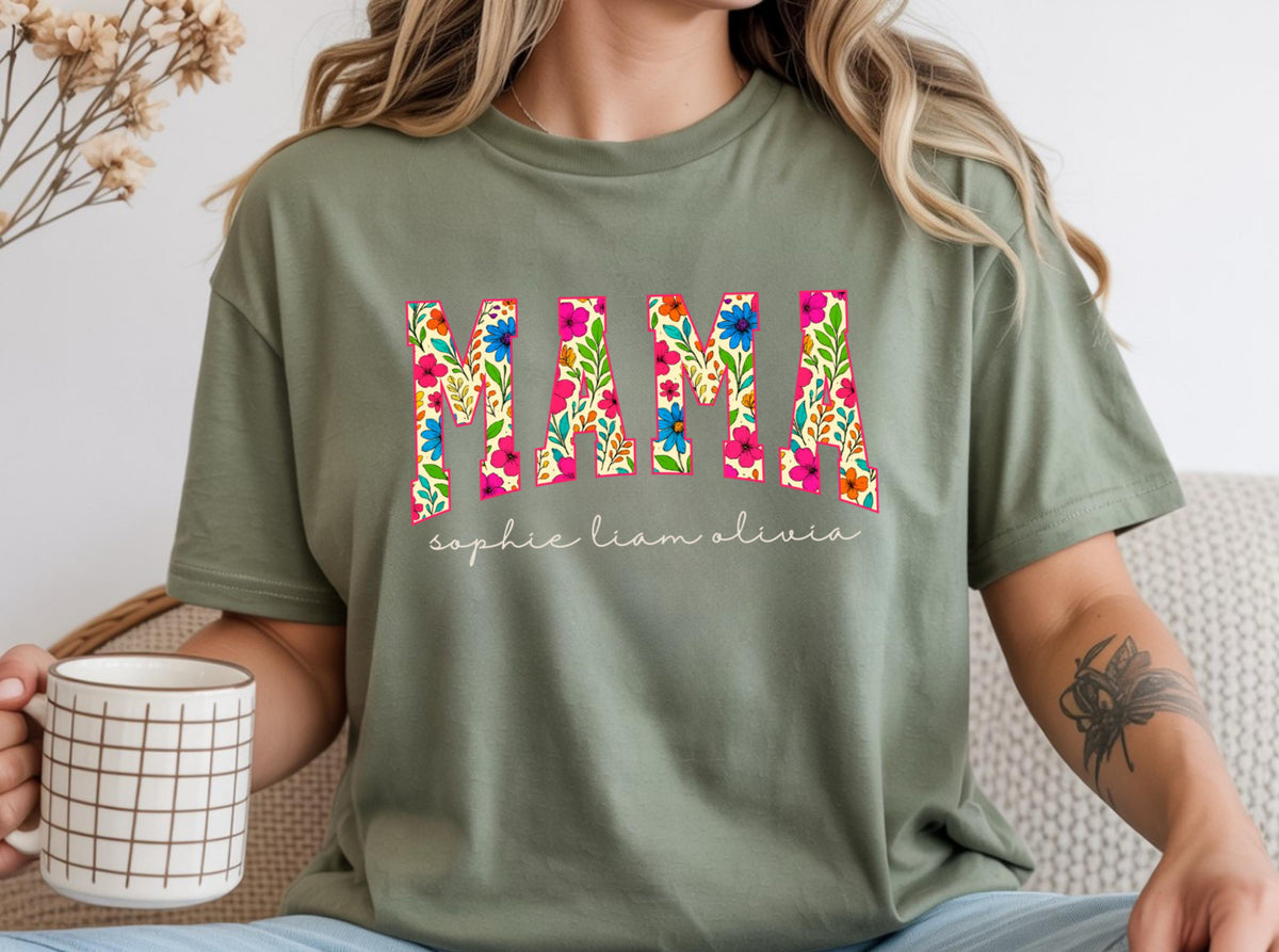 May include: Olive green t-shirt with the word "MAMA" in a floral pattern. Below the word "MAMA" are the names "sophie liam olivia" in a cursive font. The shirt is a short-sleeved, casual style.