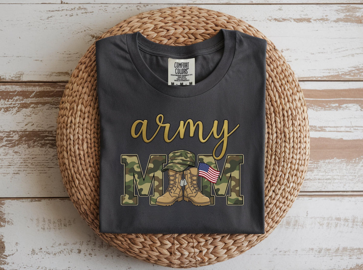 May include: Dark gray t-shirt with the words "army mom" in gold script and camouflage block letters. The design includes a military hat, boots, and an American flag. The shirt is displayed on a woven basket.