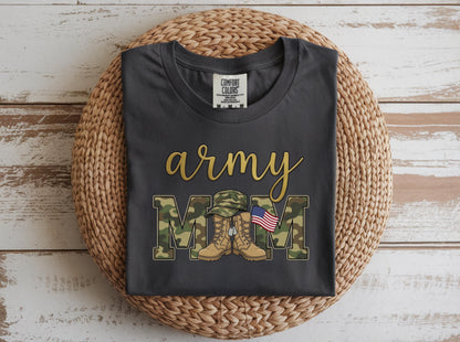 May include: Dark gray t-shirt with the words "army mom" in gold script and camouflage block letters. The design includes a military hat, boots, and an American flag. The shirt is displayed on a woven basket.