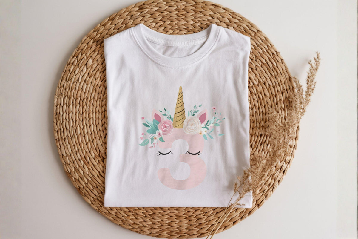 May include: White t-shirt featuring a unicorn design with a gold horn, pink ears, and floral accents. The number 3 is in pink, with closed eyes below. The shirt is displayed on a woven mat.