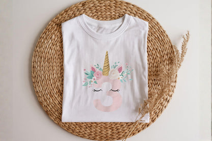 May include: White t-shirt featuring a unicorn design with a gold horn, pink ears, and floral accents. The number 3 is in pink, with closed eyes below. The shirt is displayed on a woven mat.