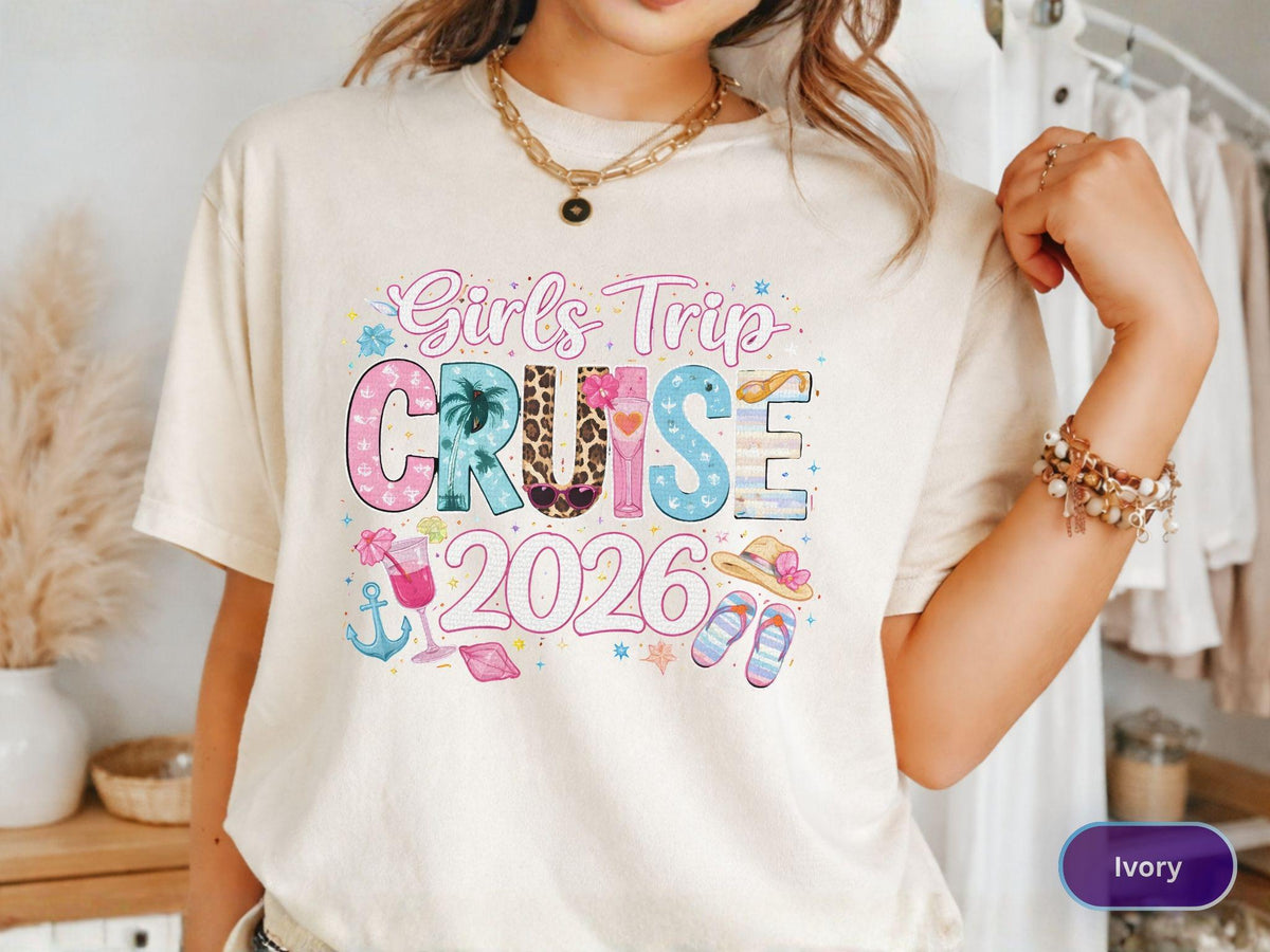 May include: Ivory-colored t-shirt with the text "Girls Trip Cruise 2026" in a colorful, playful design. The design includes images of a cocktail, anchor, sandals, and a sun hat, suggesting a vacation theme.