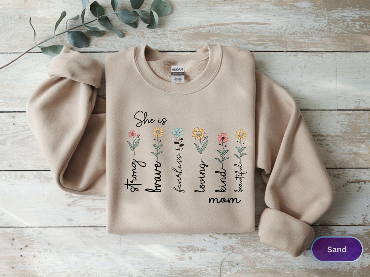 May include: A sand-colored sweatshirt with the words "She is" above a row of colorful flowers and the words "strong, brave, fearless, loving, kind, beautiful, mom" printed vertically. The sweatshirt has long sleeves and a crew neck.