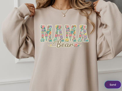 May include: Sand-colored sweatshirt with the words "MAMA Bear" in a floral design. The letters are filled with colorful flowers, and the word "Bear" is in a cursive font. The sweatshirt has a crew neck and long sleeves.