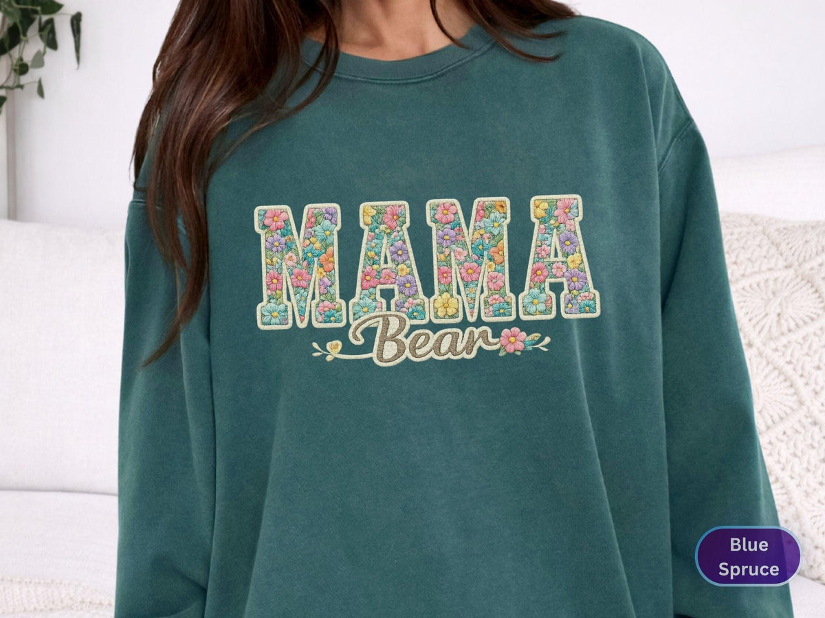 May include: Teal-colored sweatshirt with the words "MAMA Bear" in a floral design. The letters are filled with colorful flowers. The sweatshirt has a crew neck and long sleeves. The text "Blue Spruce" is in a circle in the bottom right corner.