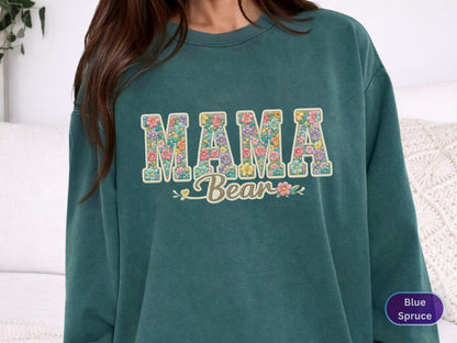 May include: Teal-colored sweatshirt with the words "MAMA Bear" in a floral design. The letters are filled with colorful flowers. The sweatshirt has a crew neck and long sleeves. The text "Blue Spruce" is in a circle in the bottom right corner.