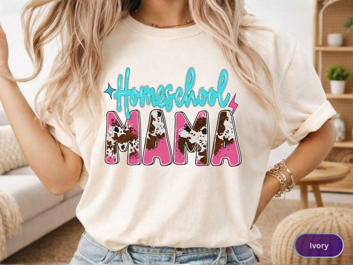 May include: Ivory-colored t-shirt with the words "Homeschool MAMA" in a turquoise script and pink block letters. The letters "MAMA" have a cow print design. The shirt has rolled-up sleeves.