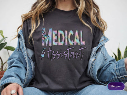 May include: Dark gray t-shirt with the words "MEDICAL ASSISTANT" in a colorful, stylized font. The word "MEDICAL" has a leopard print design. A syringe graphic is below the text. The shirt is worn with a denim jacket.