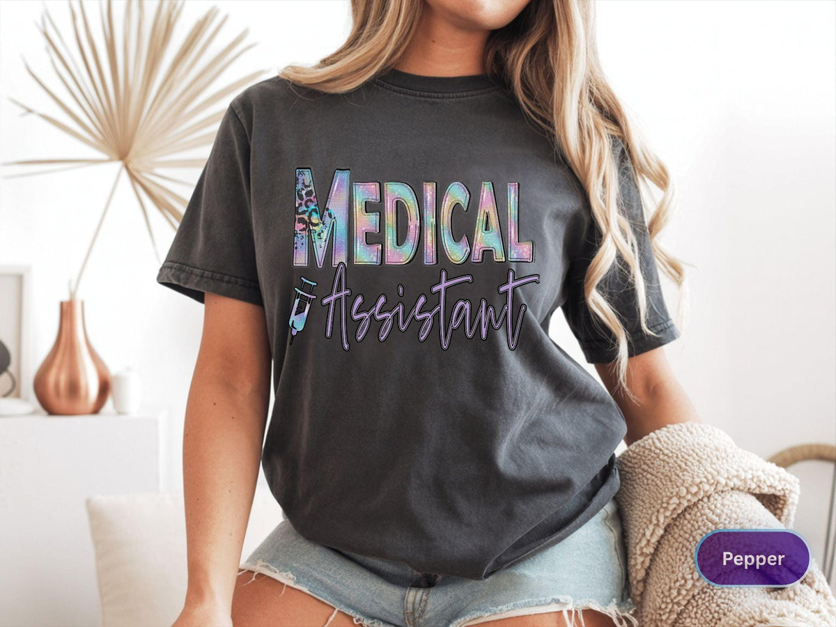 May include: Dark gray t-shirt with the words "MEDICAL ASSISTANT" in a colorful, iridescent font. The shirt features a stylized medical symbol and the word "Assistant" in a script font. The shirt is a casual style.