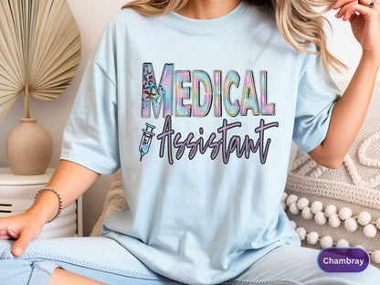 May include: Light blue t-shirt with the words "MEDICAL ASSISTANT" in a colorful, textured font. The design includes a syringe graphic. The shirt is a casual style.