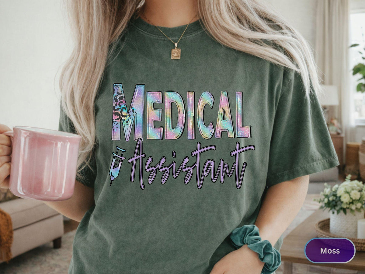 May include: A moss green t-shirt with the words "MEDICAL ASSISTANT" in a colorful, holographic font. The design includes a syringe graphic. The shirt is a short-sleeved, crew neck style.
