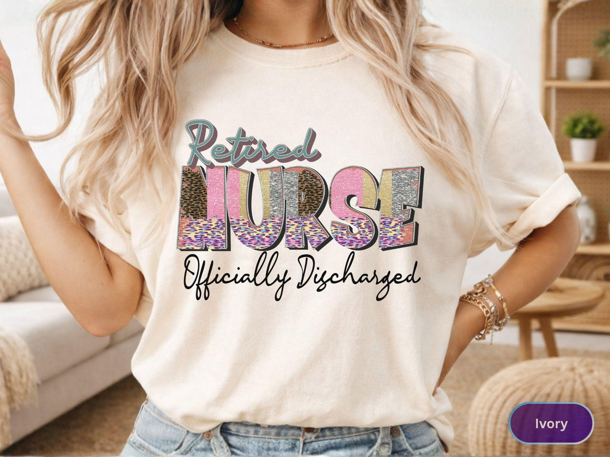 May include: Ivory-colored t-shirt with the words "Retired NURSE Officially Discharged" in a colorful, patterned design. The word "NURSE" is the focal point, with a mix of leopard print, pink, and silver glitter. The shirt is casual and comfortable.