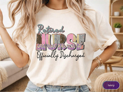 May include: Ivory-colored t-shirt with the words "Retired NURSE Officially Discharged" in a colorful, patterned design. The word "NURSE" is the focal point, with a mix of leopard print, pink, and silver glitter. The shirt is casual and comfortable.