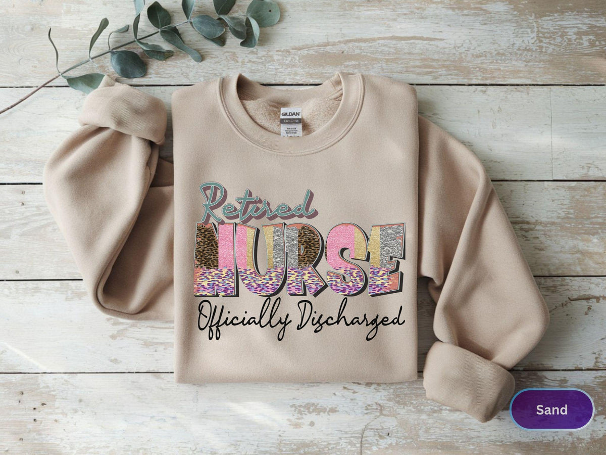 May include: A sand-colored sweatshirt with the words "Retired Nurse Officially Discharged" in a colorful, patterned design. The sweatshirt has long sleeves and a crew neck.