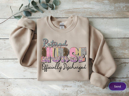 May include: A sand-colored sweatshirt with the words "Retired Nurse Officially Discharged" in a colorful, patterned design. The sweatshirt has long sleeves and a crew neck.