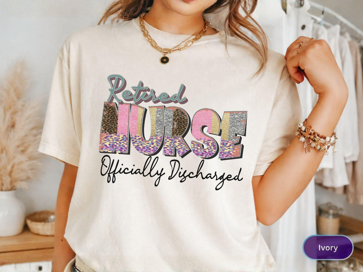 May include: Ivory-colored t-shirt with the text "Retired Nurse Officially Discharged" in a colorful, patterned design. The word "Nurse" is in pink, silver, and leopard print. The shirt has short sleeves and a crew neck.
