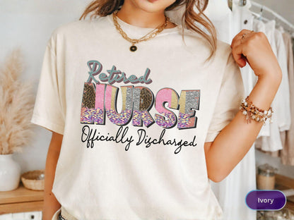 May include: Ivory-colored t-shirt with the text "Retired Nurse Officially Discharged" in a colorful, patterned design. The word "Nurse" is in pink, silver, and leopard print. The shirt has short sleeves and a crew neck.
