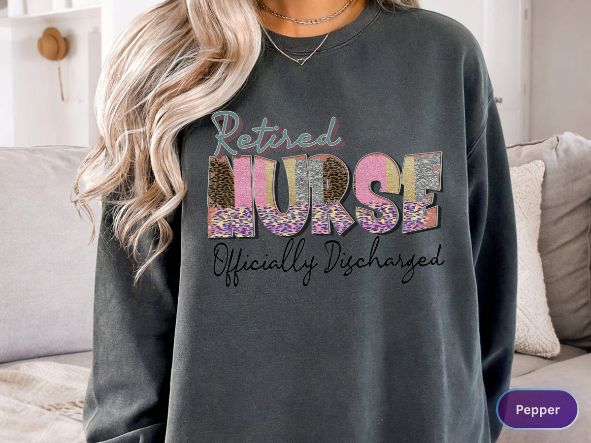 May include: Dark gray sweatshirt with the words "Retired Nurse Officially Discharged" in a colorful, patterned design. The word "NURSE" is in a mix of leopard print, pink, and gold. A comfortable, casual top.