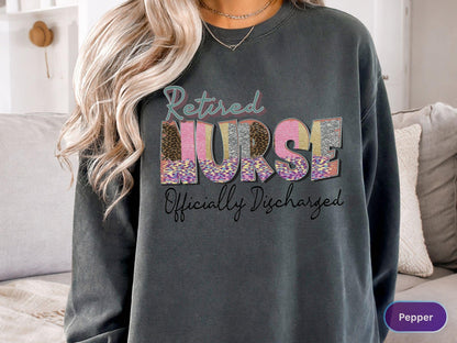 May include: Dark gray sweatshirt with the words "Retired Nurse Officially Discharged" in a colorful, patterned design. The word "NURSE" is in a mix of leopard print, pink, and gold. A comfortable, casual top.