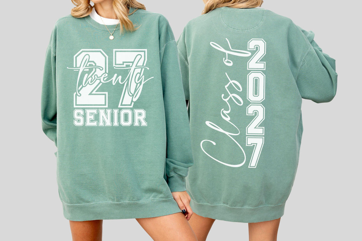 May include: A sage green sweatshirt with white lettering. The front displays "27 Twenty SENIOR" in a bold, blocky font with a script overlay. The back reads "Class of 2021" vertically in a script and block font.